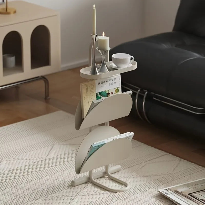 A cream-colored three-tier magazine rack styled in a modern living room setting next to a black leather sofa, holding magazines, a candle, and a teacup.