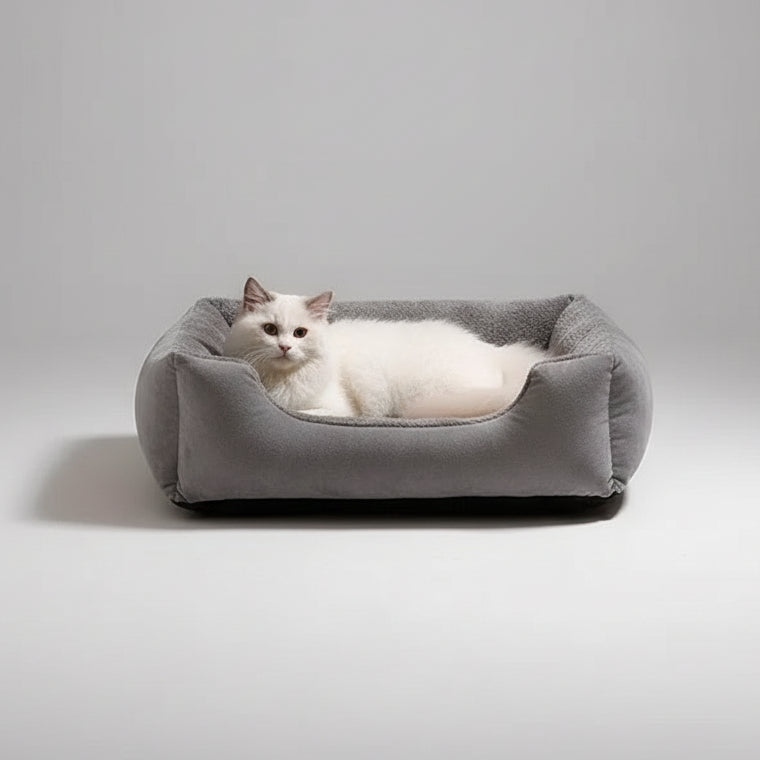 A fluffy white cat rests inside a square gray plush pet bed on a cream shag rug, with part of another pet bed visible in the background.