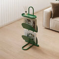A green metal magazine rack with three curved shelves and a top hanger, holding two magazines in a bright living room with light wood floors and a cream sofa.