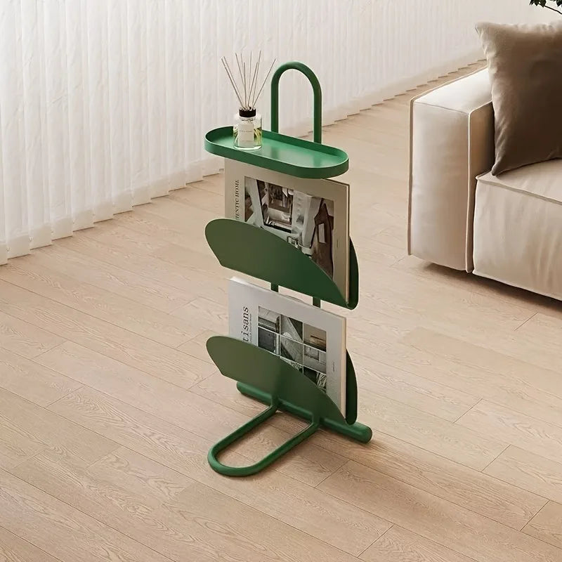 A green metal magazine rack with three curved shelves and a top hanger, holding two magazines in a bright living room with light wood floors and a cream sofa.