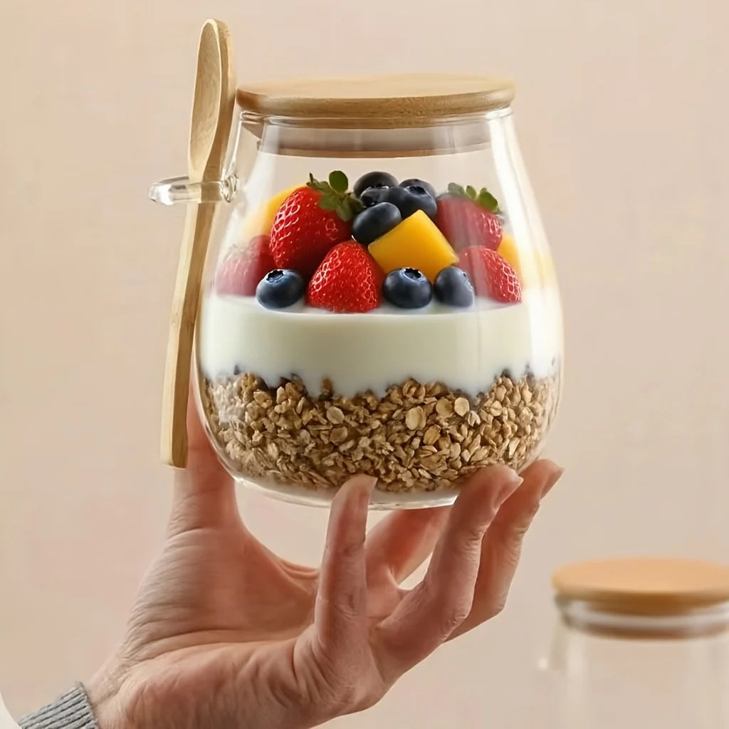 A hand holds a clear glass jar with a bamboo lid and side handle, filled with granola, yogurt, and fresh fruit including strawberries, blueberries, and mango pieces. A small bamboo spoon is attached to the side.