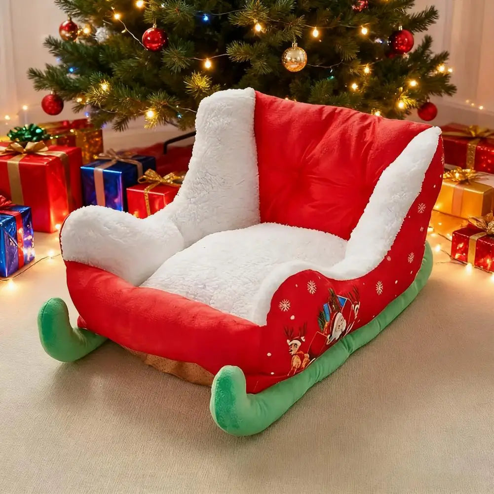A plush Christmas-themed pet chair designed to look like a sleigh, featuring a red and white faux-fur interior, a red exterior with green sleigh-style wheels, and holiday illustrations on the side, set in front of a decorated Christmas tree with wrapped gifts.
