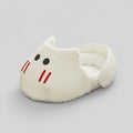 A plush white cat-shaped slipper or small pet bed with embroidered black eyes and red whiskers, placed on a geometric patterned rug in a room setting.