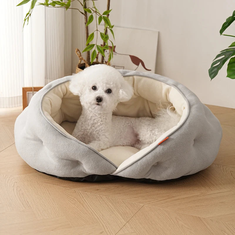 A small white dog sits inside a round, plush pet bed with a light gray exterior and cream interior, placed on a light wood floor in a bright room.