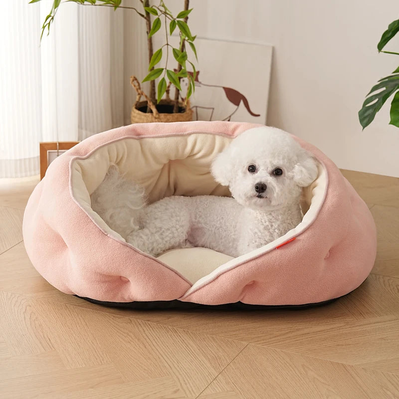 A small white dog sits inside a round, plush pink pet bed with a cream-colored lining, placed on a light wood floor in a bright room.