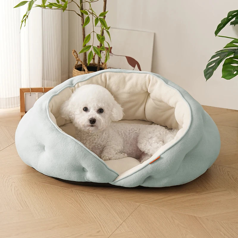 A small white dog sits inside a round, tufted pet bed with a pale mint green exterior and cream interior, placed on a light wood floor in a bright room with plants in the background.
