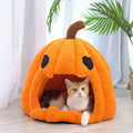 An orange pumpkin-shaped cat bed with a plush fuzzy texture, black embroidered eyes, and a brown stem, with a cat sitting inside on a light wood floor in front of white curtains.