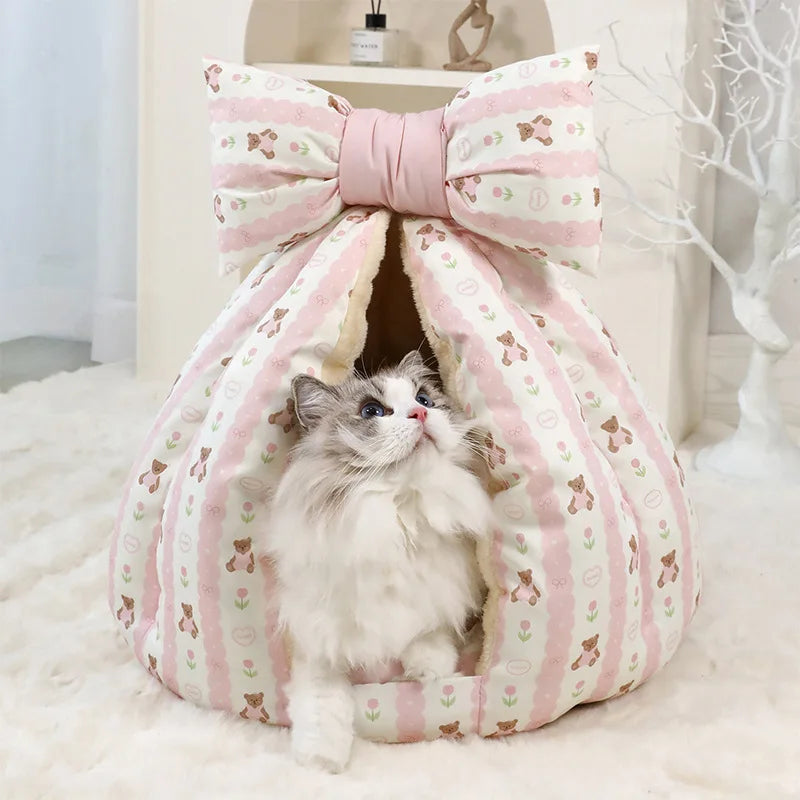 Cat sitting inside a pink pet bed with a large bow on top, on a soft white surface.