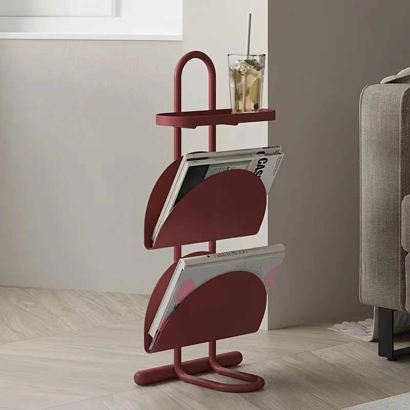 Red magazine rack with books and a glass on a wooden floor next to a gray sofa.