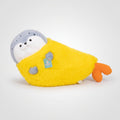 A plush penguin toy wrapped in a bright yellow blanket, resting on a beige sofa in a softly lit room. The penguin has a gray head, white belly, and orange feet, with a small blue hangtag attached.
