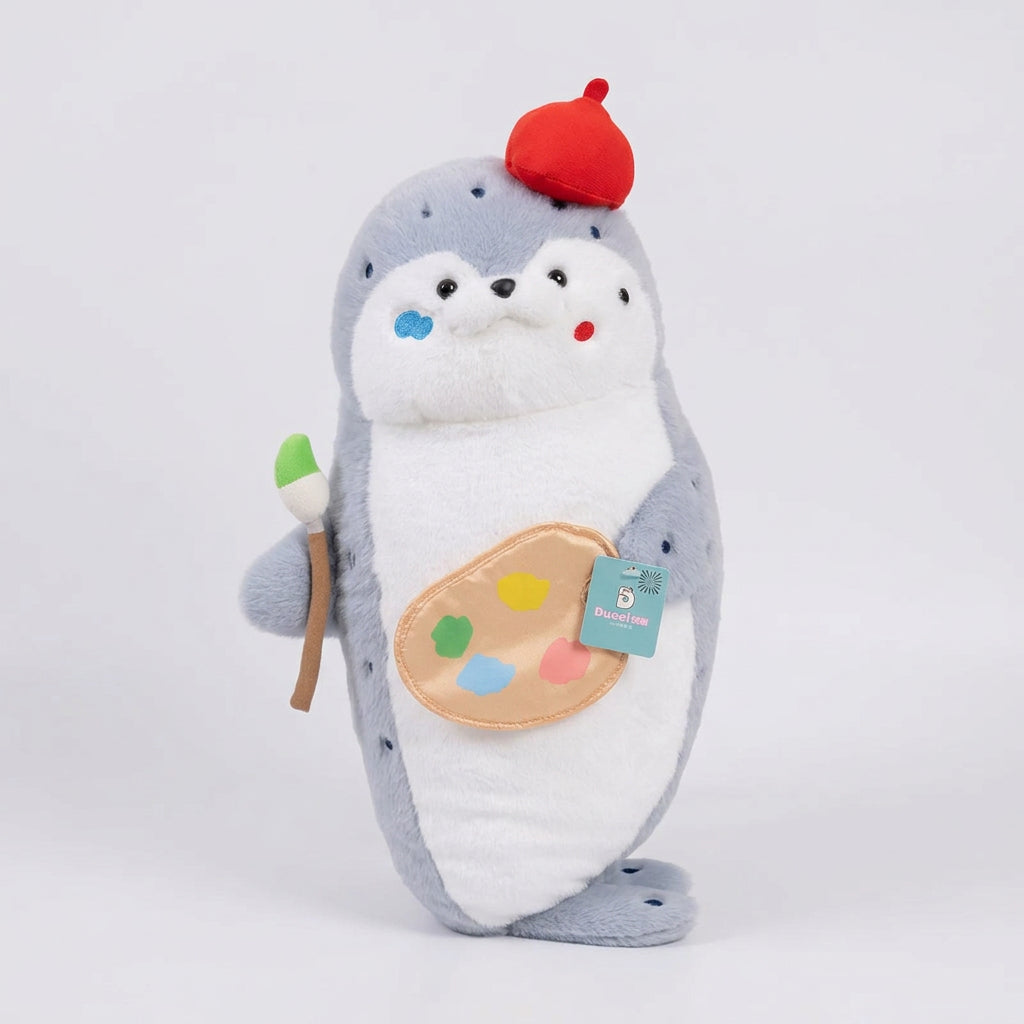 A plush seal pillow styled as an artist, resting on a beige armchair in a bright room. The plush seal is gray and white with a red beret, holding a paintbrush, and has a paint palette patch on its belly.