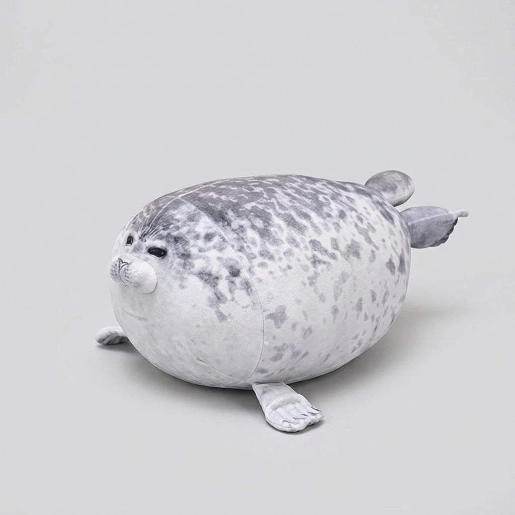 A plush seal toy with a white and gray spotted pattern, lying on a wooden shelf against a dark green wall with a leafy plant and a bottle in the background.