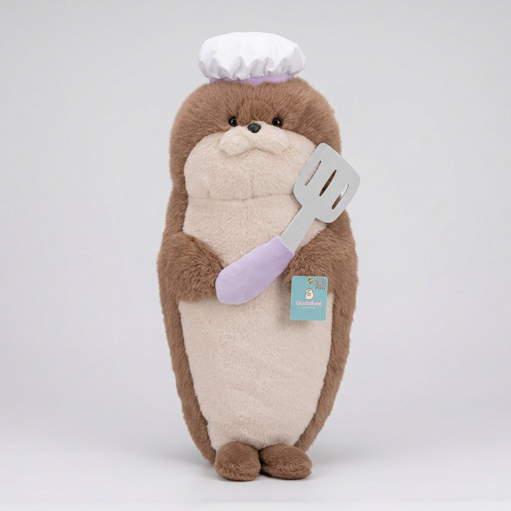 A plush bear toy styled as a baker, wearing a white chef's hat and holding a white spatula with a lavender handle, sitting on a cream-colored armchair in a bright room.